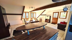 A dining area with a large wooden table, chairs, and a ceiling fan. There are windows with curtains, a clock on the wall, and a door with a decorative handle.