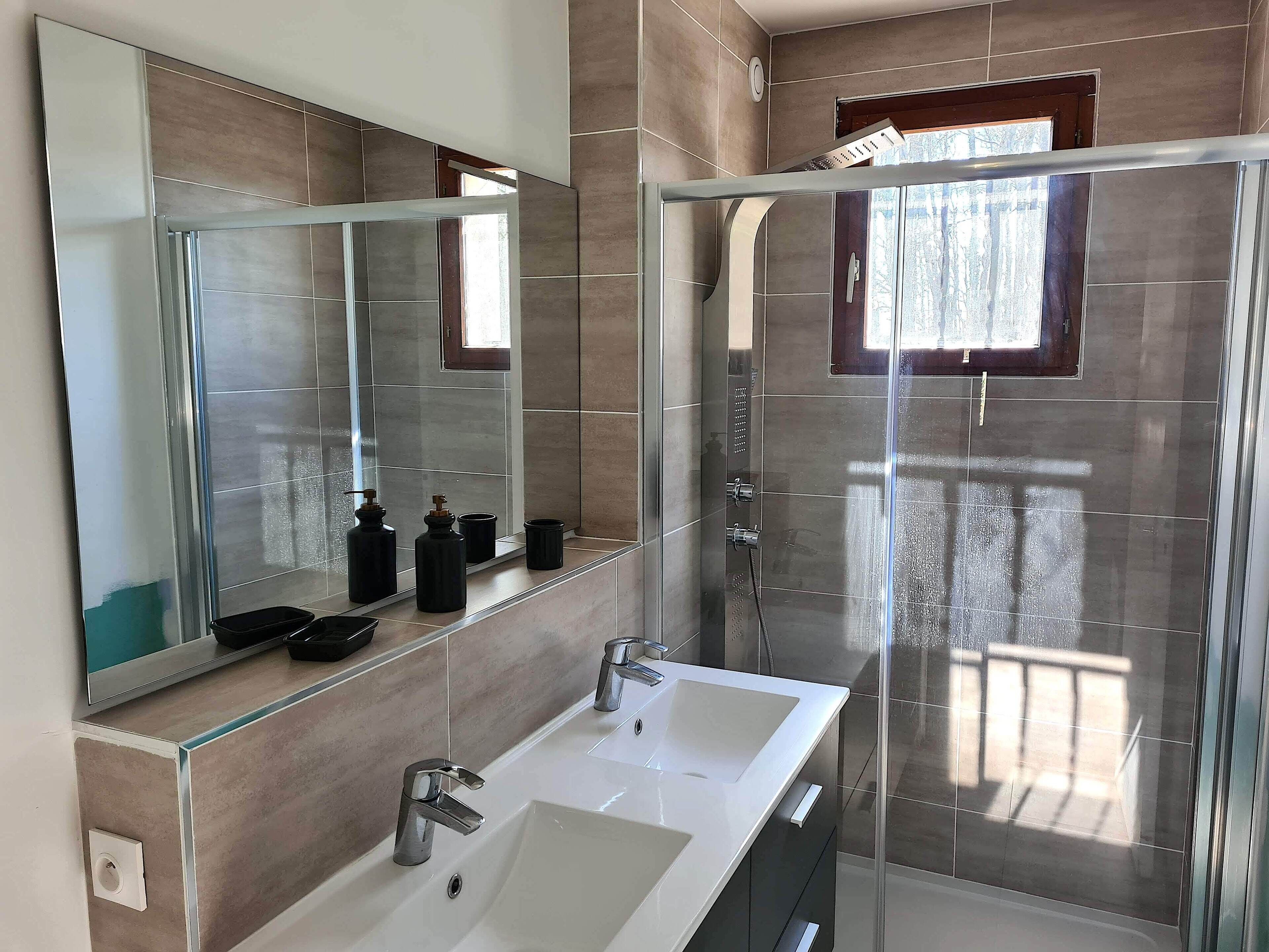 A modern bathroom with a large mirror, two sinks, a shower enclosure, and a window.