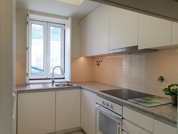 A modern kitchen with white cabinets, a stainless steel sink, a built-in oven, and a window with a view of the outside. - Station House (Guimarães)