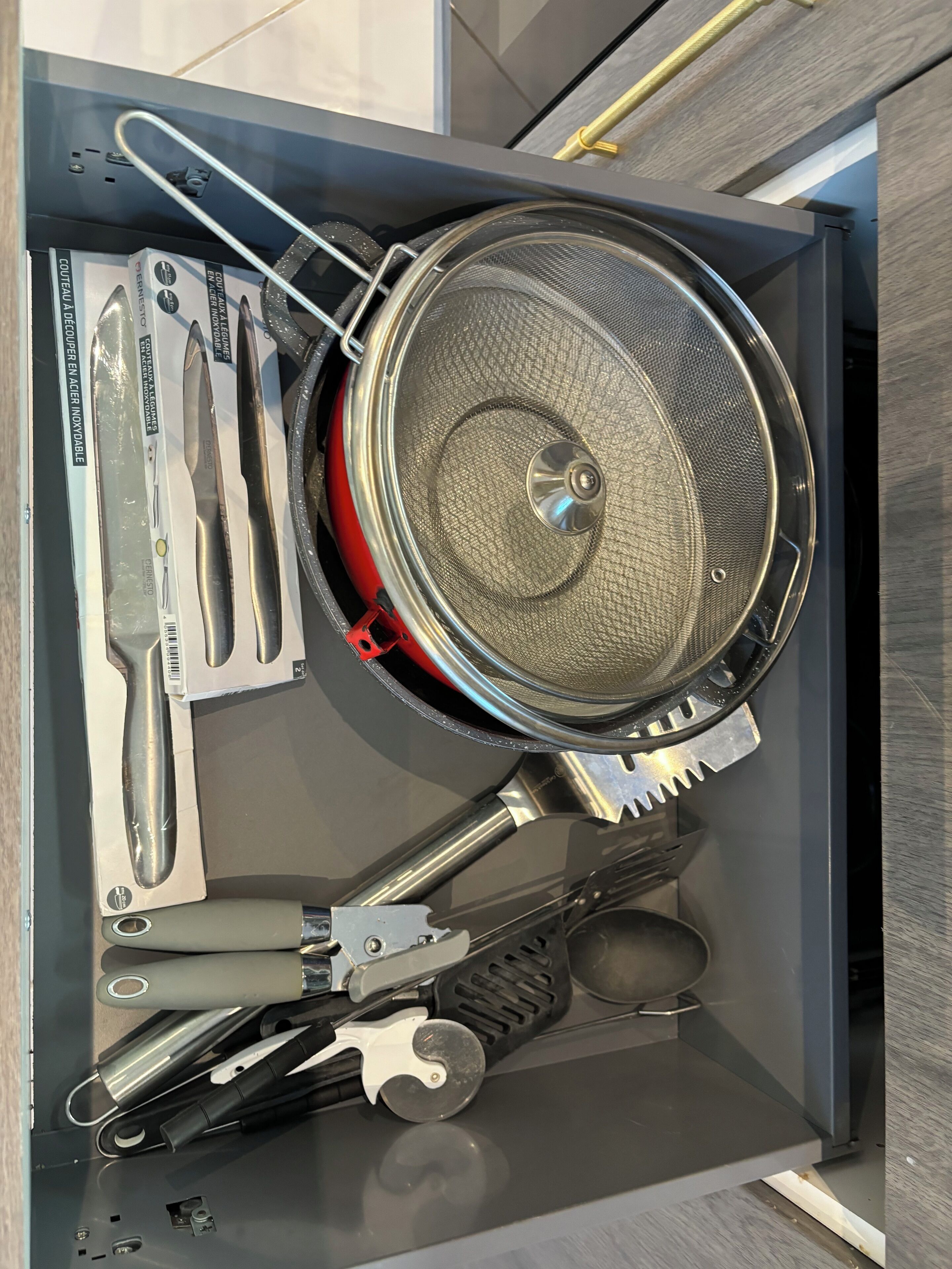 Un cajón con utensilios de cocina, como un colador, un cuchillo y un abrelatas.