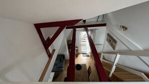 A view from a high vantage point within a building, looking down a staircase with red beams. The interior includes a wooden floor, a small table, and a bench.