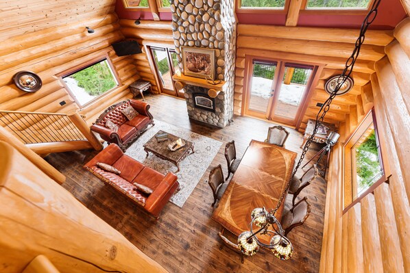 A log cabin interior with a stone fireplace, wooden furniture, and large windows. - Luxury log house in Sunshine Valley “Ravenloft” (Hope)