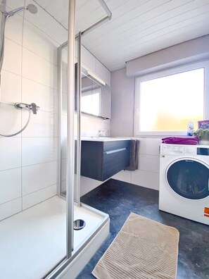A modern bathroom with a glass shower enclosure, a built-in sink, a washing machine, and a window providing natural light. - Beautiful, modern lakeside apartment—the perfect place to relax and unwind (Immenstaad am Bodensee)