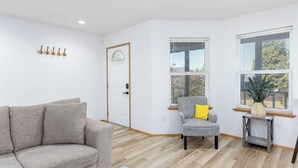 A modern living room with a grey sofa, a small armchair with a yellow pillow, and a wooden coffee table. There are two windows with blinds, a door with a glass panel, and a wall-mounted coat rack.