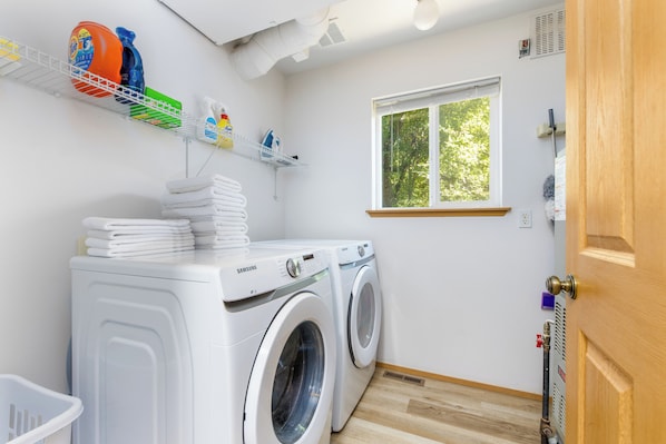 A laundry room with two white Samsung washing machines, a drying rack, and a window with a view of trees.