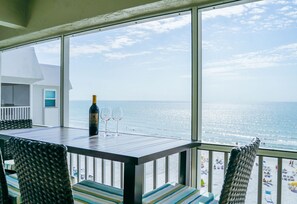 Uma varanda com uma mesa, duas cadeiras, uma garrafa de vinho e duas taças. A vista inclui o oceano e uma praia com pessoas e guarda-sóis.