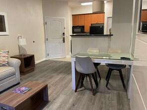 A compact living space with a kitchenette, a glass dining table, two chairs, a sofa, a TV on a wooden stand, and a door. - Suite · The Point Lux Suite, close to Universal (Orlando)
