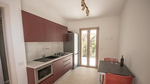 A compact kitchen with a refrigerator, stove, and sink. There is a dining table with chairs and a bottle of wine. - North Shell (Cala Gonone)