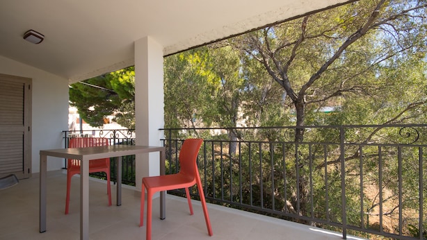 A balcony with a table and two chairs, overlooking a garden with trees and a fence. - North Shell (Cala Gonone)