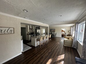 A spacious living area with a dark wood floor, a dining area with a long table and chairs, a kitchen with white cabinets and stainless steel appliances, and a large window with patterned curtains.