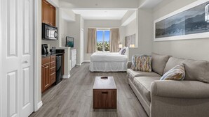 A compact hotel room with a bed, a sofa, a small table, a kitchenette, and a large window with curtains. - Studio · The point Studio 10 min Universal (Orlando)