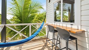 Een houten terras met een hangmat, een bar met een metalen stoel, en uitzicht op palmbomen.