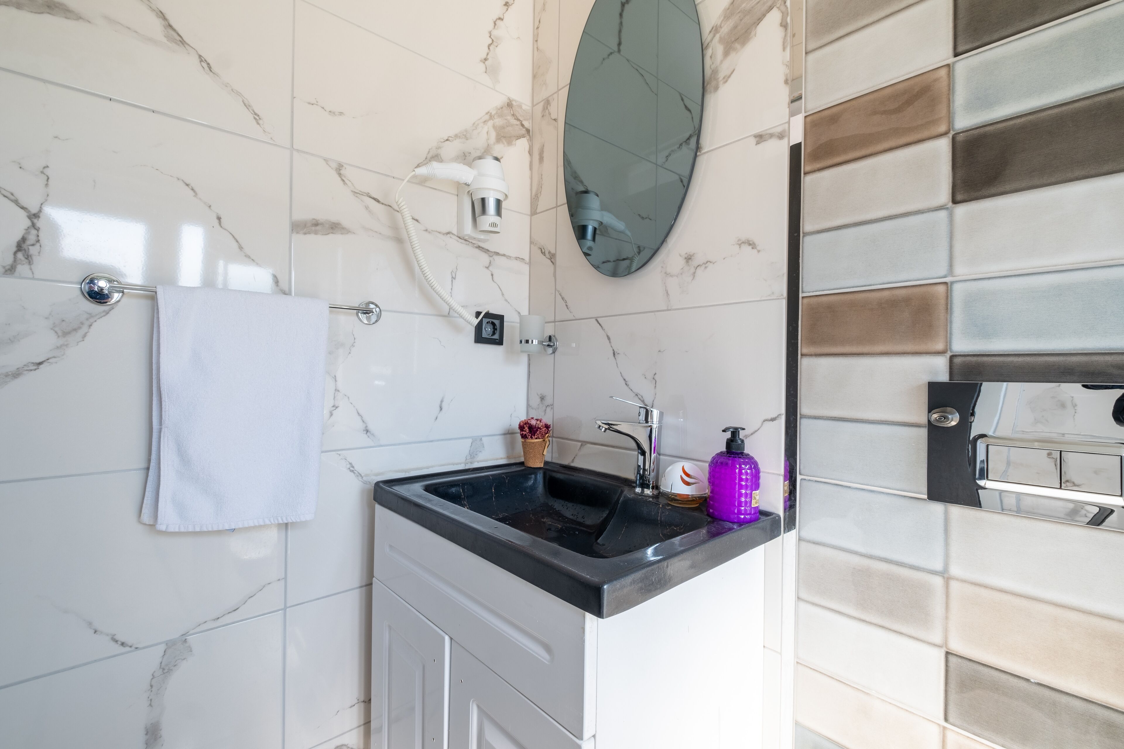Une salle de bain avec un mur en marbre, un lavabo doté d’un plan de travail noir, une serviette blanche, une petite plante en pot et un miroir.