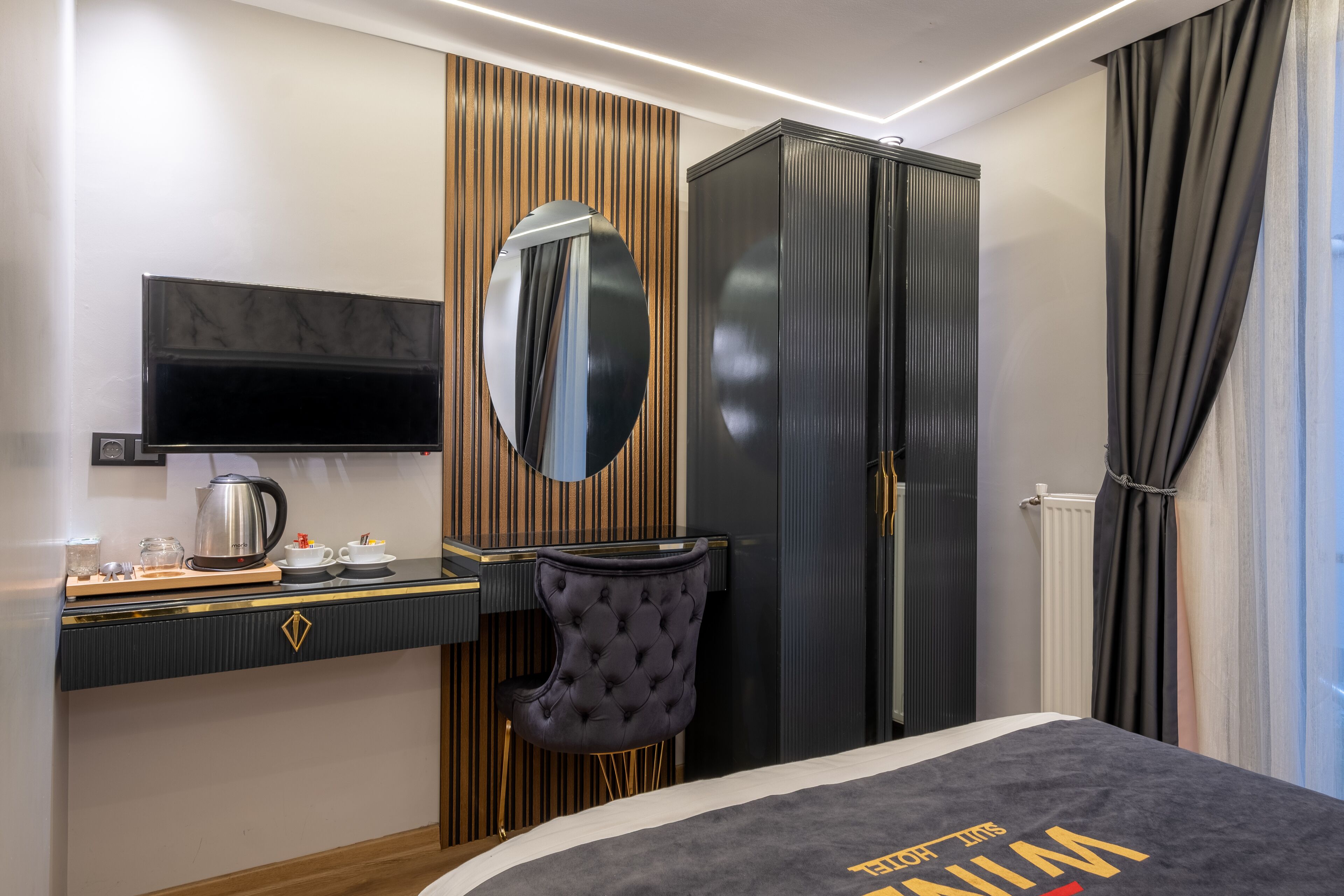 A modern hotel room with a bed, a desk with a coffee maker, a chair, a wardrobe, and a television.