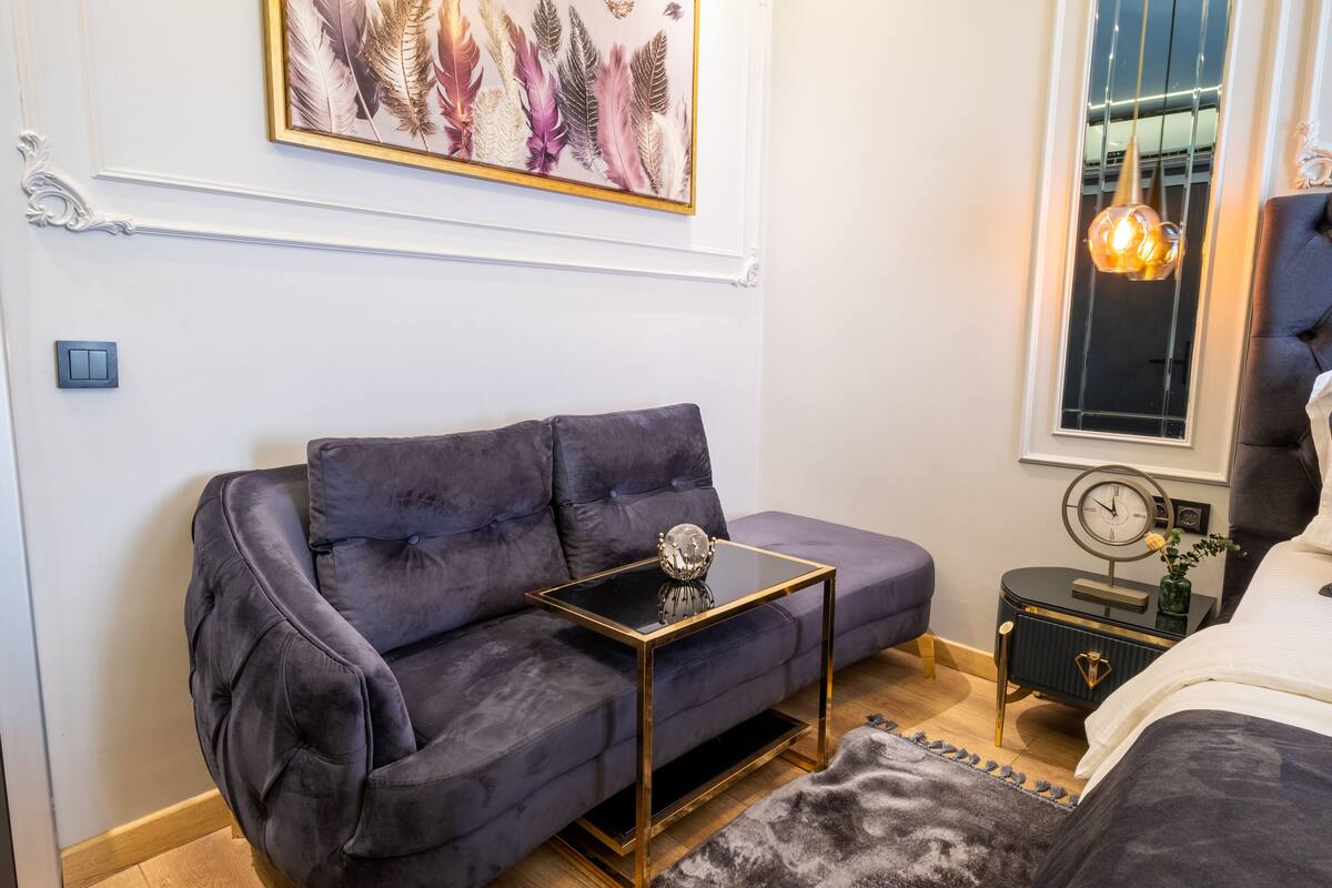 A modern bedroom with a dark grey sofa, a glass-top side table, a small bedside table with a clock, a bed with a white duvet, and a window with a hanging light fixture.