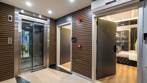 Un hall d’hôtel moderne avec un ascenseur en verre, un mur recouvert de panneaux en bois et un panneau indiquant le numéro de la chambre 302.
