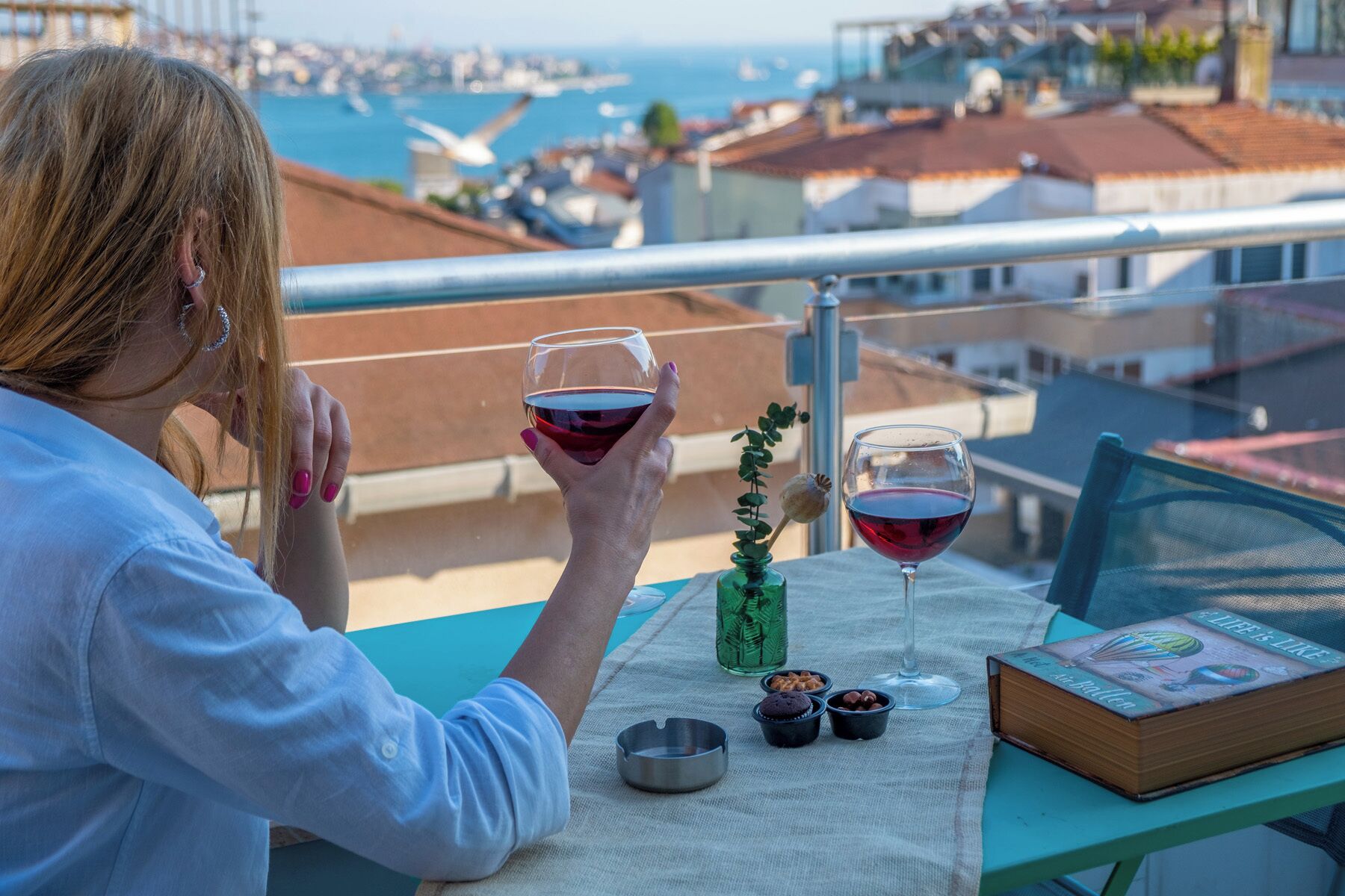 Una persona sentada en una mesa con un libro, dos copas de vino tinto, una pequeña planta y algunos bocadillos, en un balcón con vistas a un puerto deportivo y edificios.