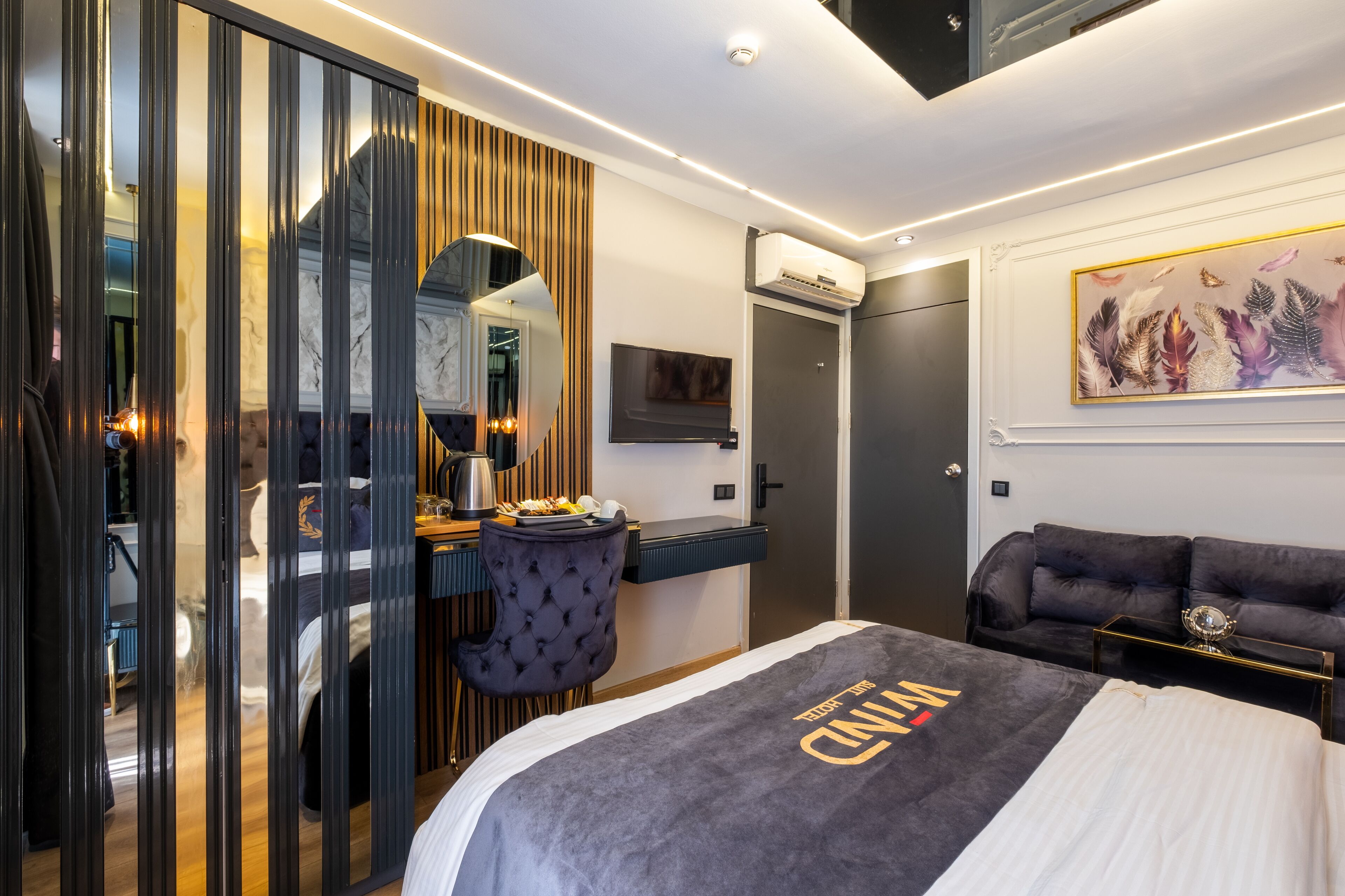 A modern hotel room with a bed, a desk, a chair, a sofa, a mirror, an air conditioning unit, and a painting on the wall.