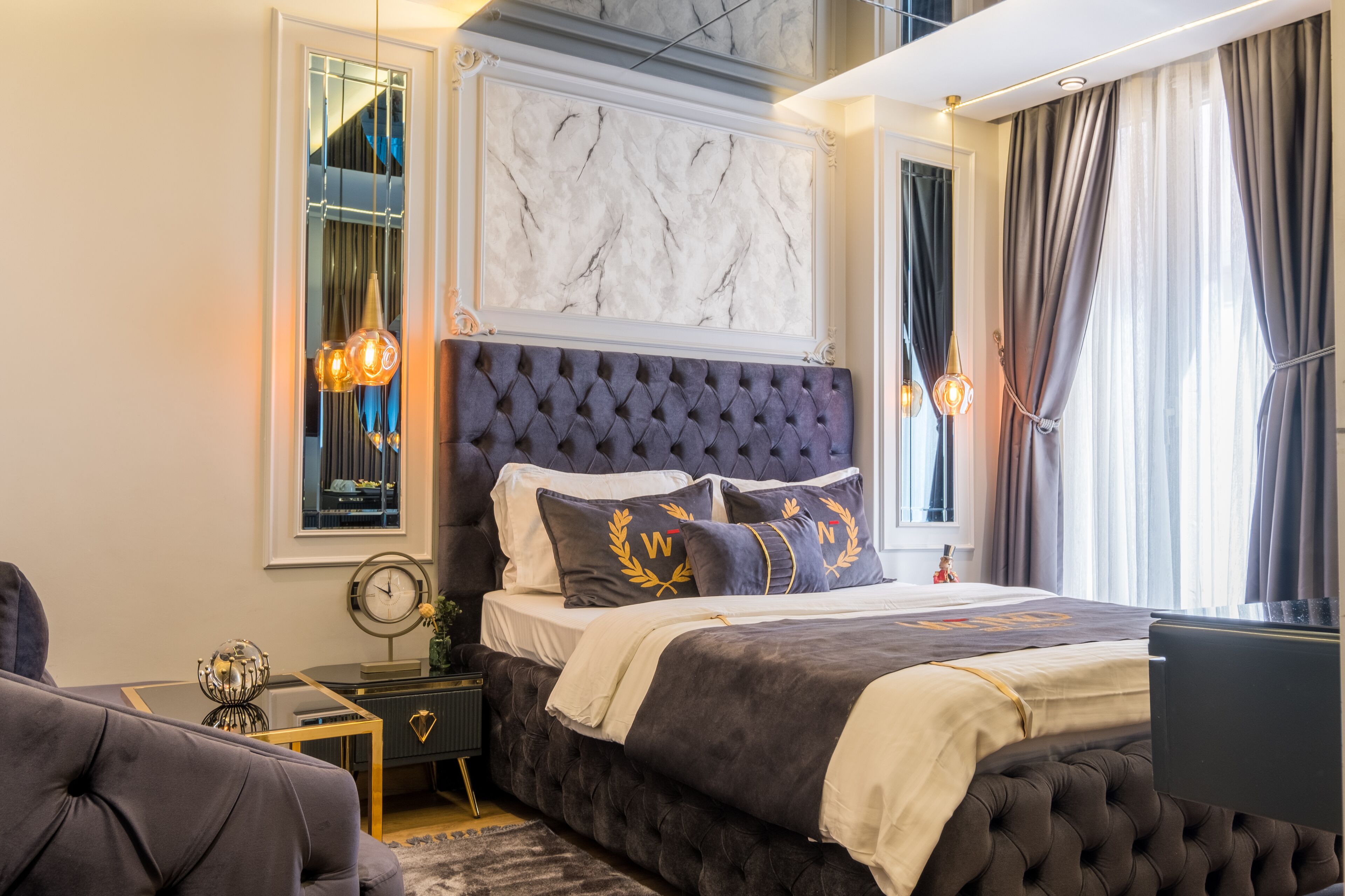 A bedroom with a tufted headboard, a large mirror, and a bed with a dark-colored comforter and decorative pillows.