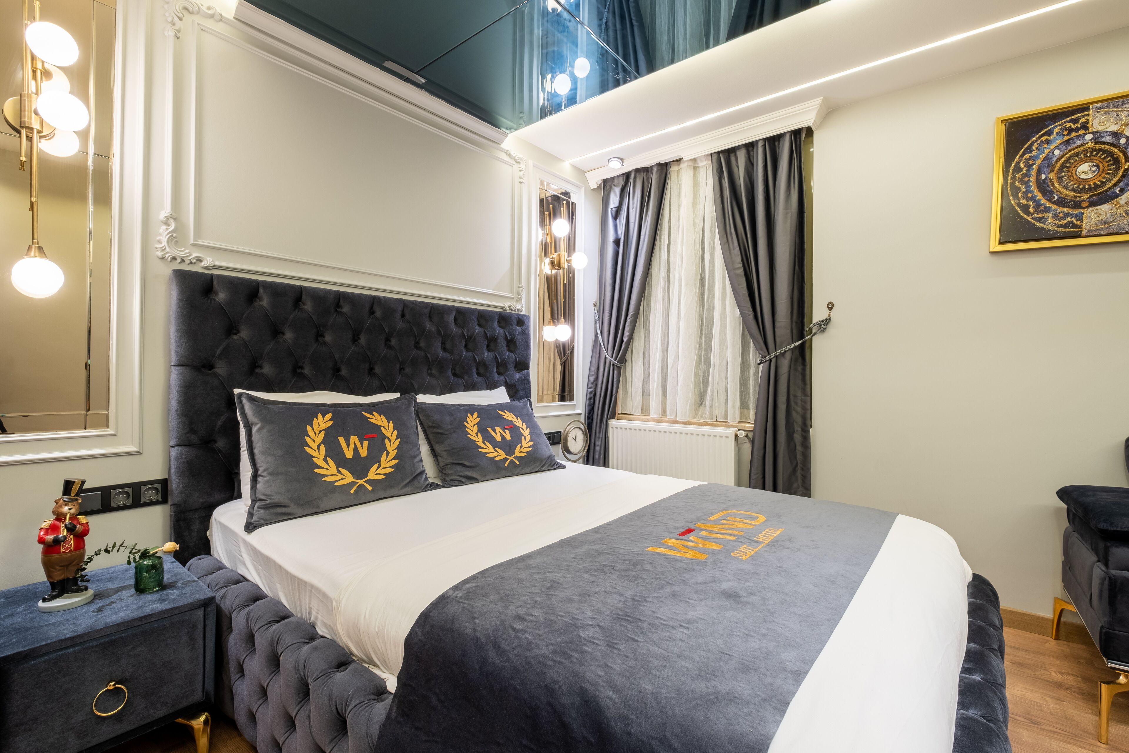 Une chambre à coucher avec une tête de lit capitonnée, un lit recouvert d’une couvre-lit grise et blanche, et un coussin décoratif orné d’un motif de couronne de laurier.
