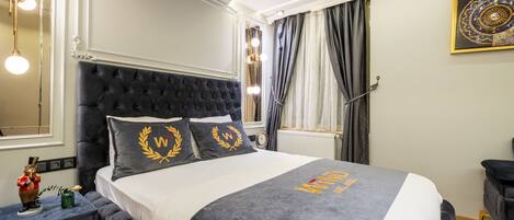 Une chambre à coucher avec une tête de lit capitonnée, un lit recouvert d’une couvre-lit grise et blanche, et un coussin décoratif orné d’un motif de couronne de laurier.