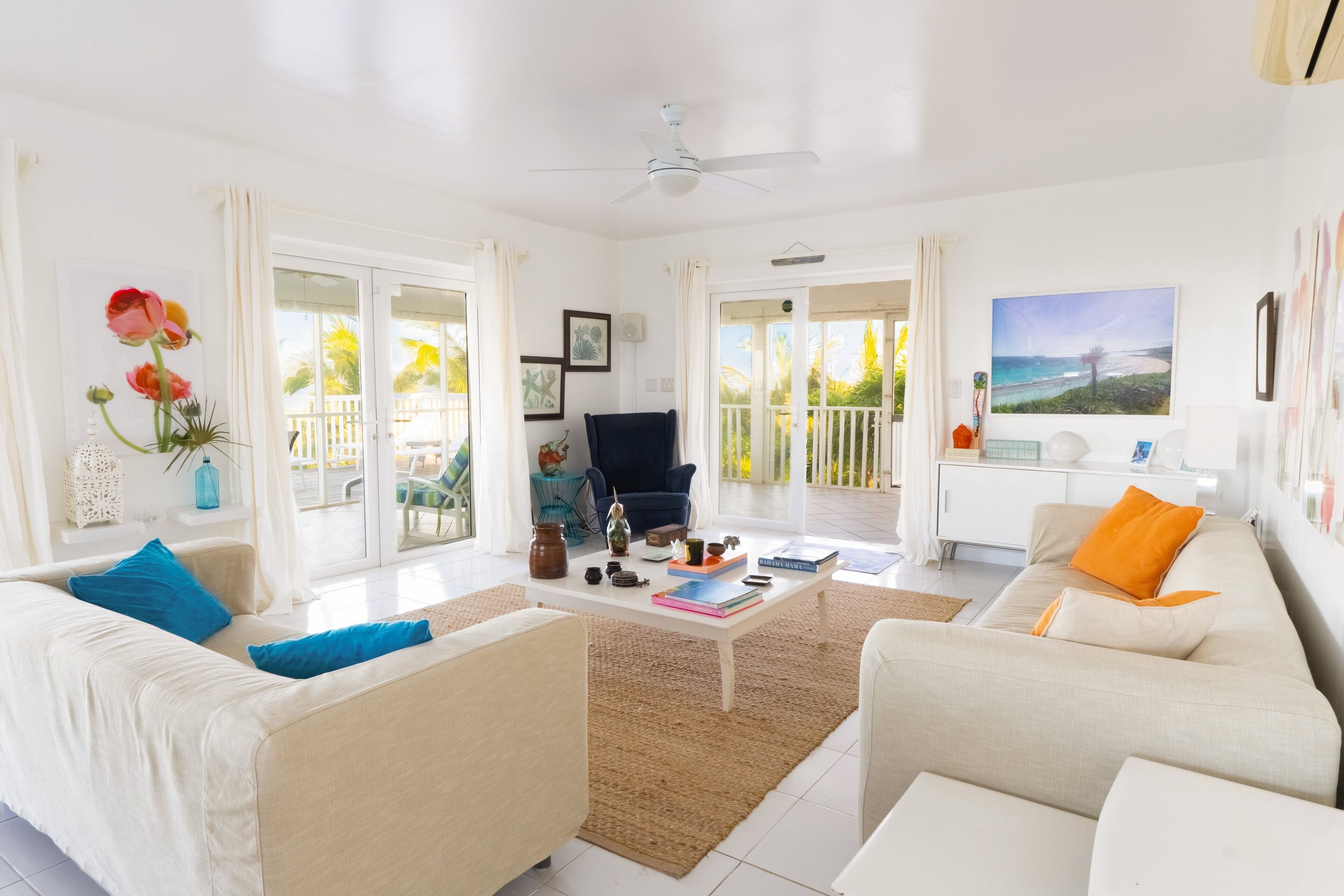 A bright living room with a white sofa, a coffee table, and a TV. There are curtains, a ceiling fan, and a painting on the wall.