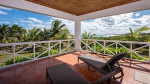 A covered outdoor area with a wooden ceiling, white railings, and a view of palm trees and the ocean.