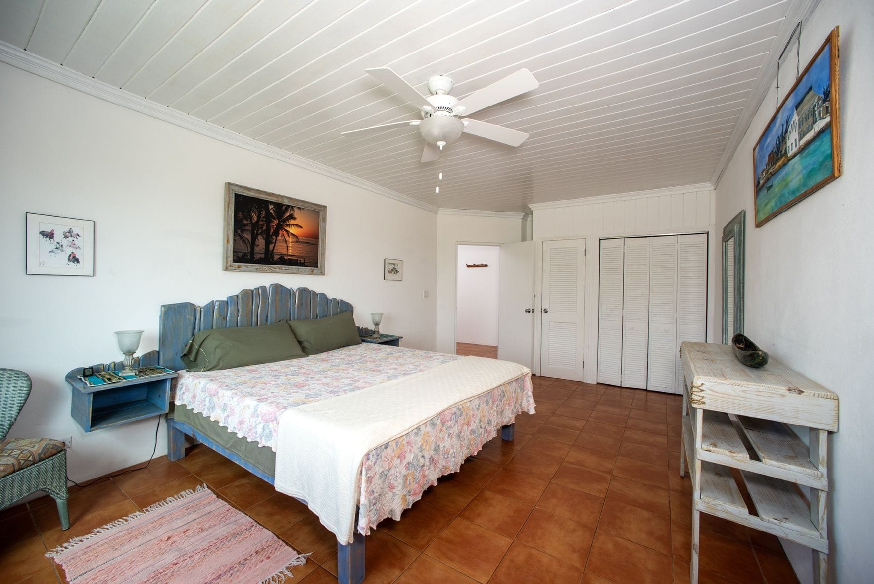 Quarto com cama, ventoinha no teto, mesa de cabeceira, penteadeira e porta.