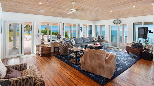 A spacious living room with a large sofa, a coffee table, and a TV. There are multiple windows offering a view of the ocean.
