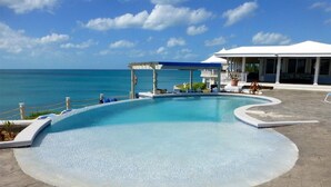 A pool with a curved edge, a covered seating area, and a view of the ocean.