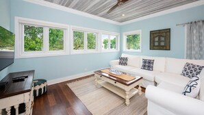 Una sala de estar con un sofá blanco, una mesa de centro y un televisor. Hay ventanas amplias con vistas al verdor exterior.