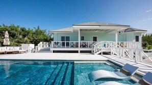 Un área junto a la piscina con una piscina azul cristalina, sillones blancos para tomar el sol y una terraza cubierta.
