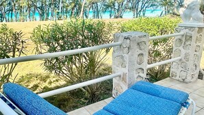 A sun lounger with a blue cushion on a tiled patio overlooking a beach with palm trees and a clear sky. - Moonflower 4 Bedroom Home (Gregory Town)