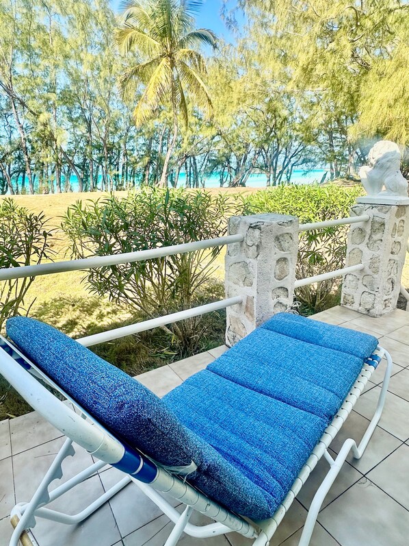 A sun lounger with a blue cushion on a tiled patio overlooking a beach with palm trees and a clear sky.
