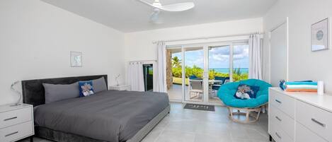 A modern bedroom with a bed, a ceiling fan, a balcony with a chair, and a view of the ocean.