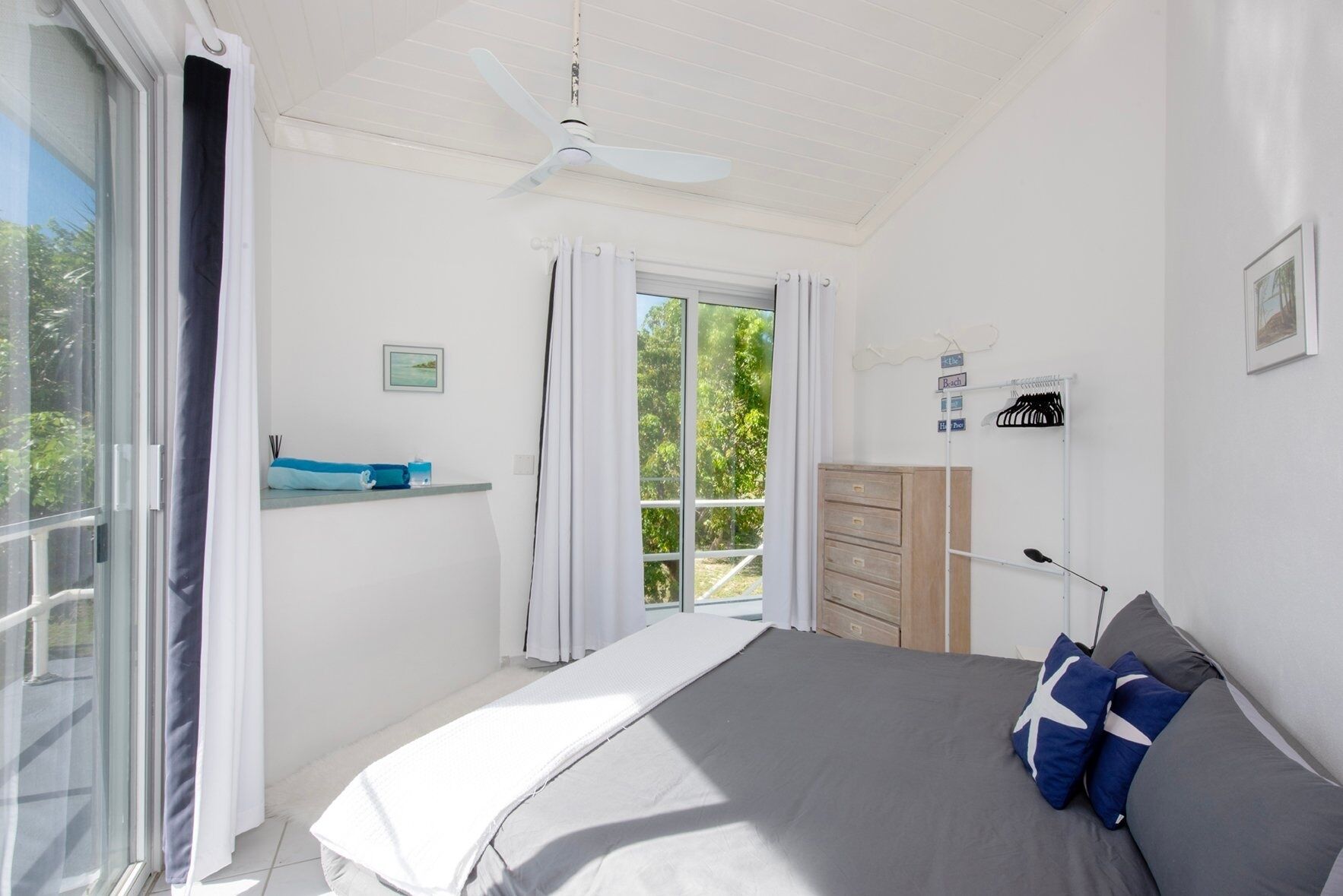 Un dormitorio con cama, cómoda, ventilador de techo, ventana con cortinas y vista a zonas verdes.