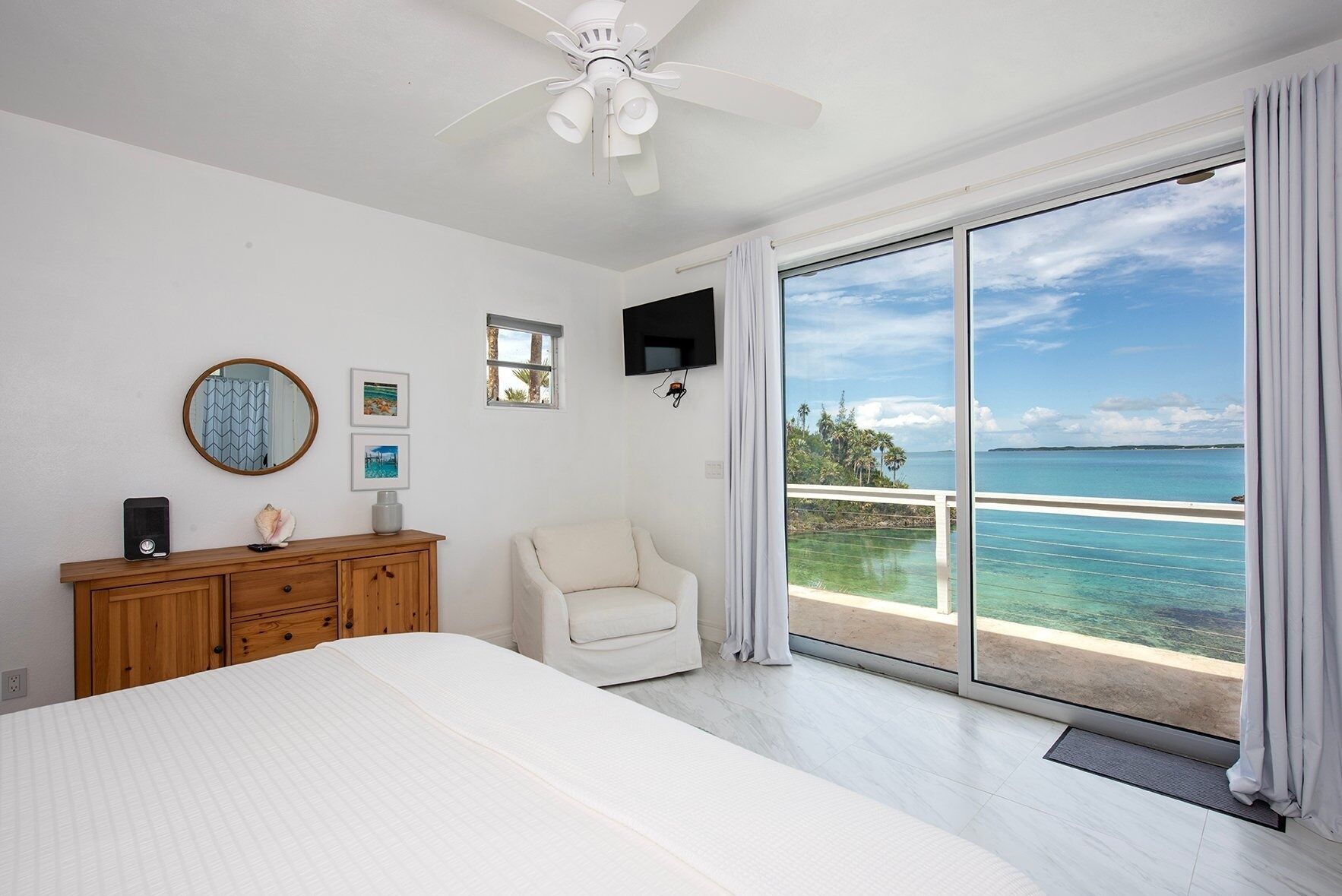A bedroom with a bed, a wooden dresser, a white armchair, a ceiling fan, a TV, and a large sliding glass door leading to a balcony with a view of the ocean.