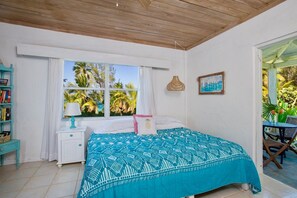 A bedroom with a bed, a nightstand, a window with a view of palm trees, a wicker lamp, and a painting on the wall. - Poponi House 2 Bedroom Home (North Palmetto Point)