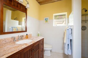 A bathroom with a marble sink, wooden cabinets, a white toilet, a shower, and a window with a view. - Poponi House 2 Bedroom Home (North Palmetto Point)