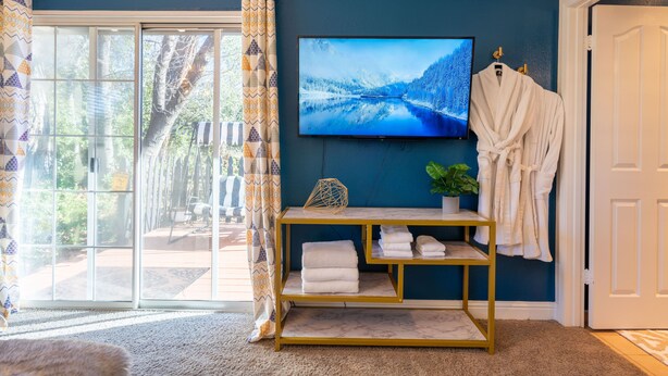 Una habitación con un televisor grande en la pared, una puerta corrediza de vidrio, una mesita de consola con marco dorado y toallas, y una bata colgada en la pared.