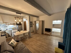 A modern living room with a wooden floor, a sofa, a dining table with chairs, a TV, and a kitchen area with a fridge, microwave, and hanging lights. - Neaspace - 19 Rue Neuve (Nice)