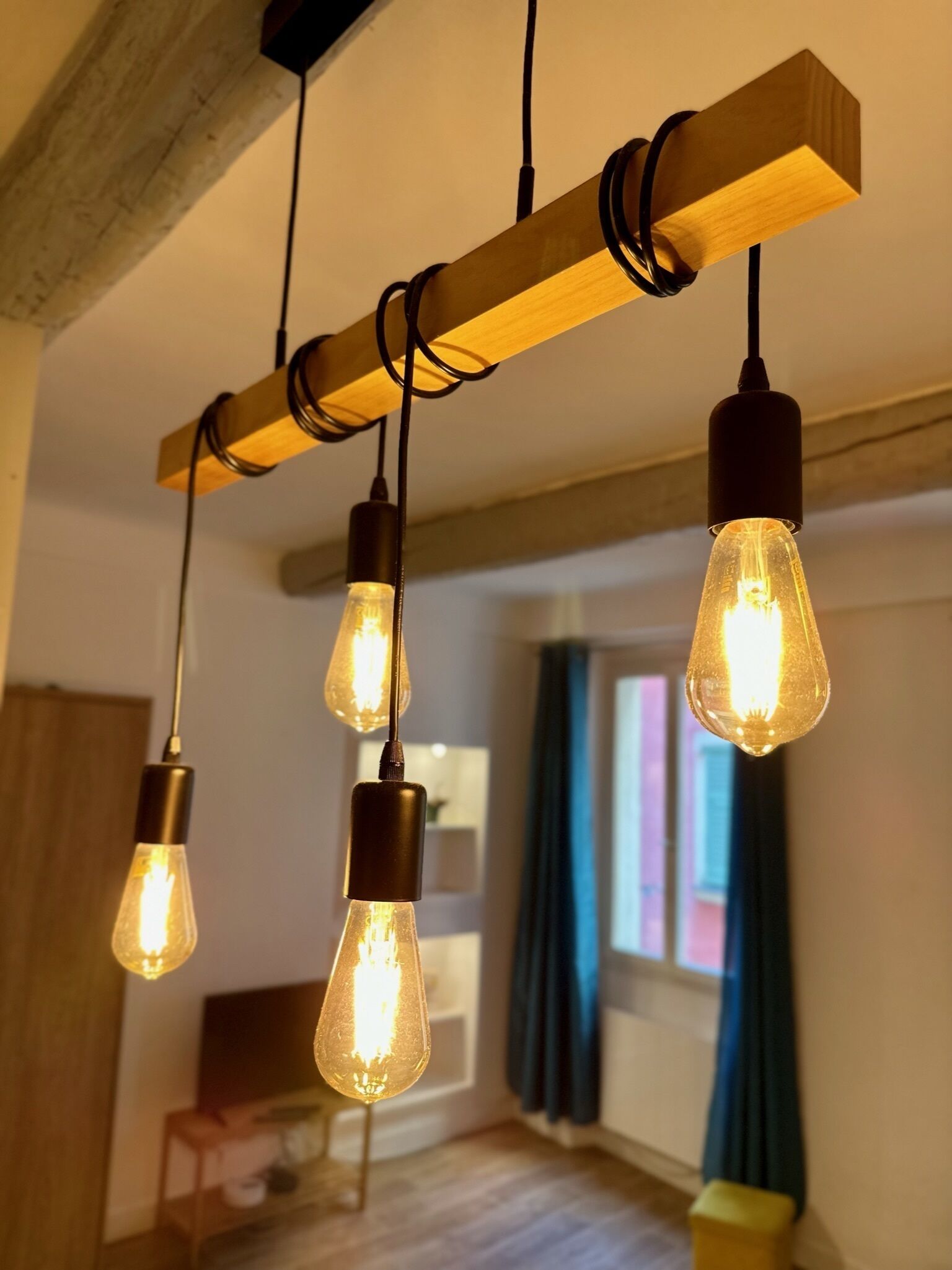 En modern inredning med synliga träbjälkar, hängande Edison-lampor och ett fönster med gardiner.