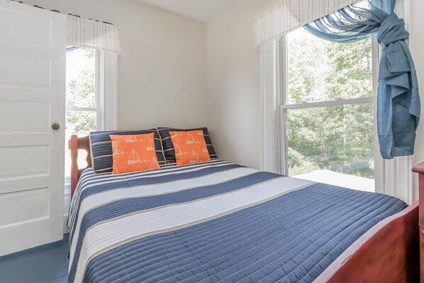 A bedroom with a bed, striped bedding, two pillows with sailboat patterns, and a view of trees through the window. - Updated Long Island Maine Cottage (Long Island)