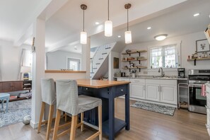 A modern kitchen with a central island, wooden chairs, pendant lights, and a staircase leading to an upper level. - Updated Long Island Maine Cottage (Long Island)