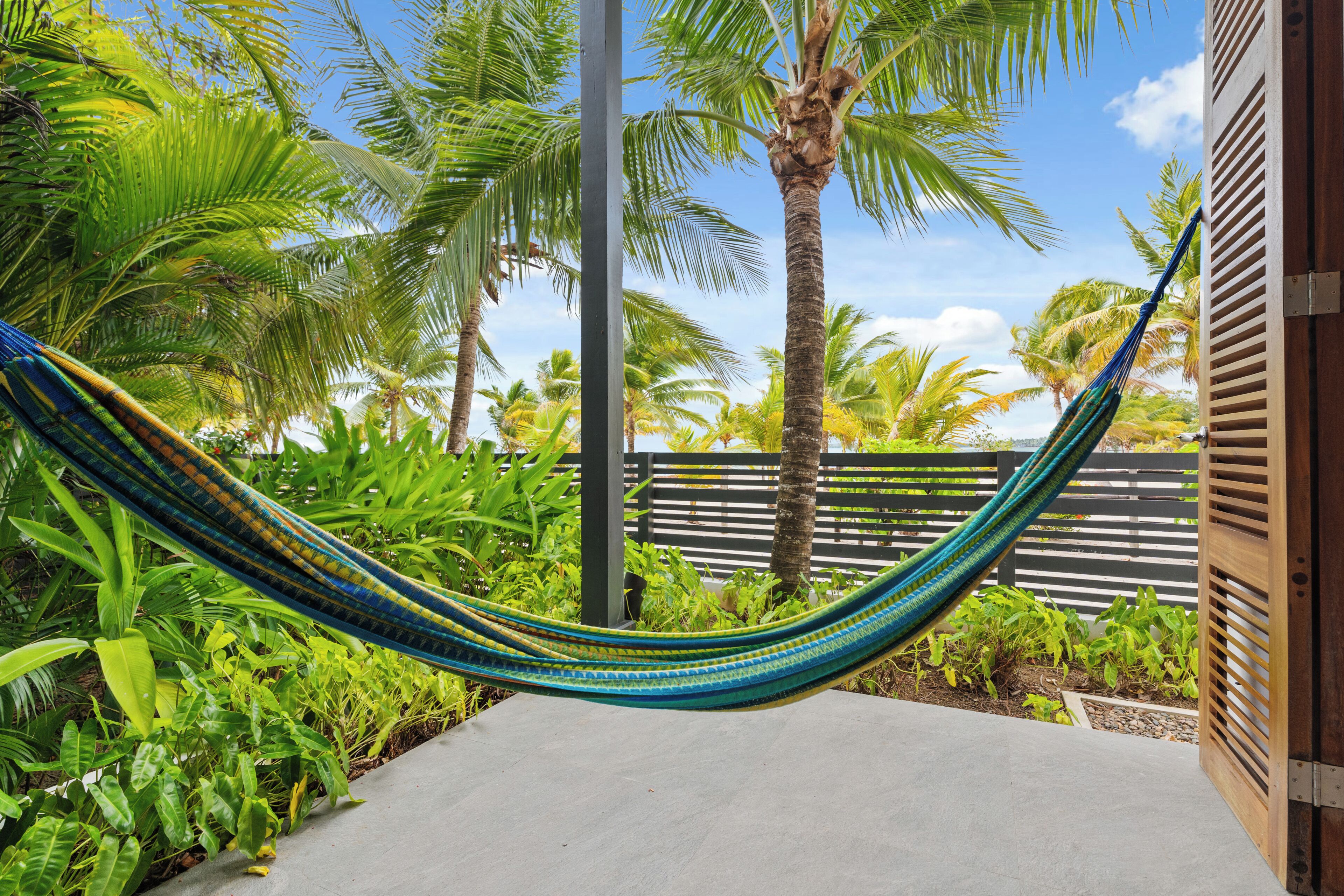 A hammock in a tropical garden with palm trees and a wooden fence.