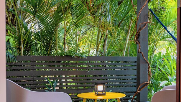 A patio area with a yellow table and two chairs, surrounded by lush green foliage and a wooden fence.