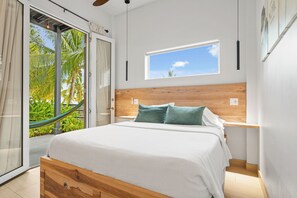 A bedroom with a large bed, a wooden headboard, a window with a view of palm trees, and a hammock outside the door.