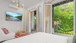 A modern bedroom with a large bed, a TV on the wall, and a view of lush greenery through the open door.
