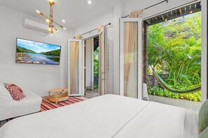 A modern bedroom with a large bed, a TV on the wall, and a view of lush greenery through the open door.
