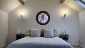 Quarto com cama grande, duas mesas de cabeceira, uma janela redonda e teto inclinado com vigas de madeira.